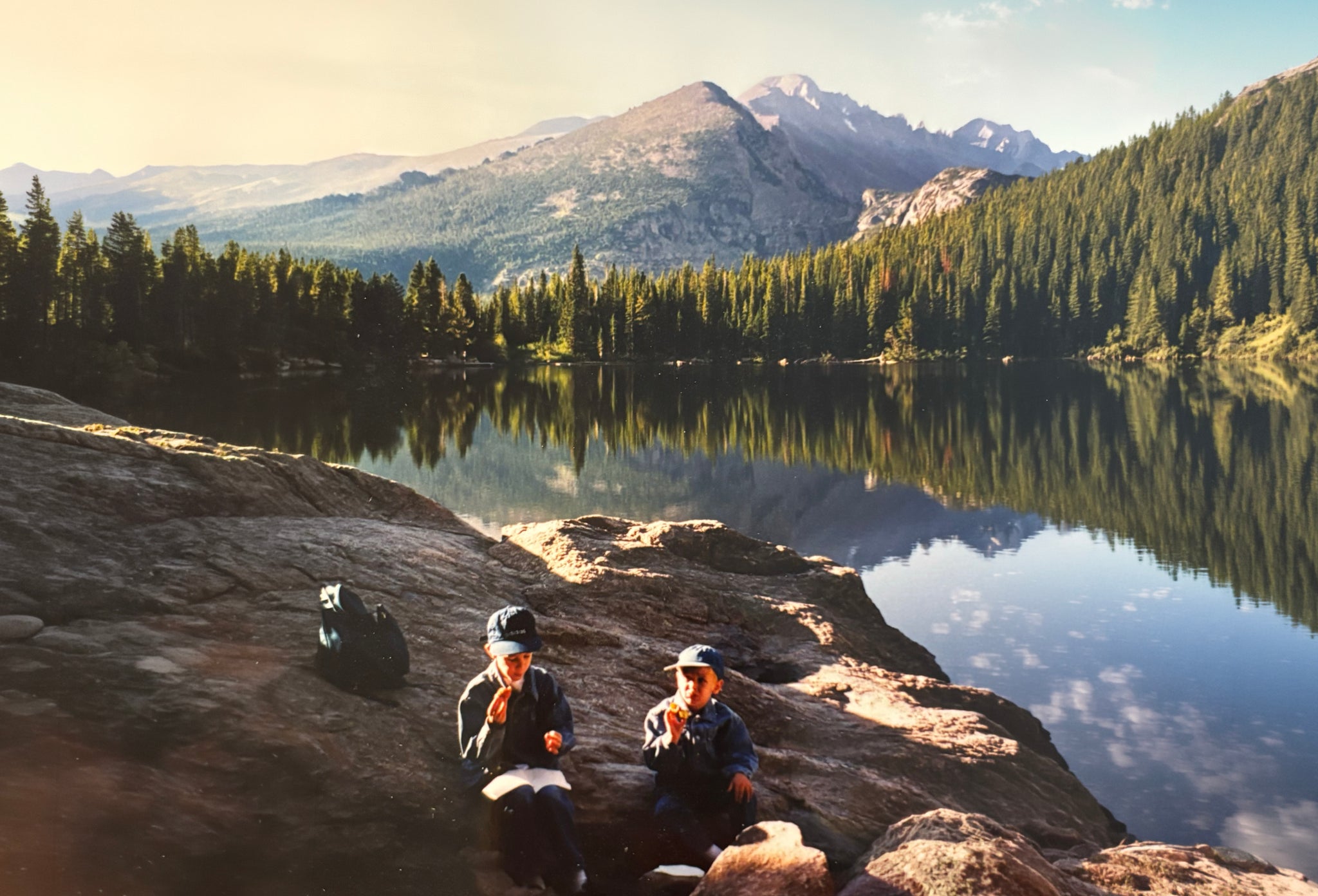 Two brothers as kids on a rock by an alpine lake, snacks in hand, mountains behind them.