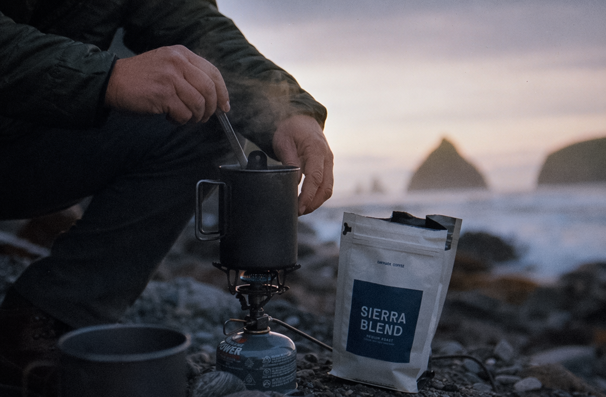 A person brews Daymark Coffee's Sierra Blend on a backpacking stove at a rocky coastal beach at dusk, with sea stacks visible in the background.