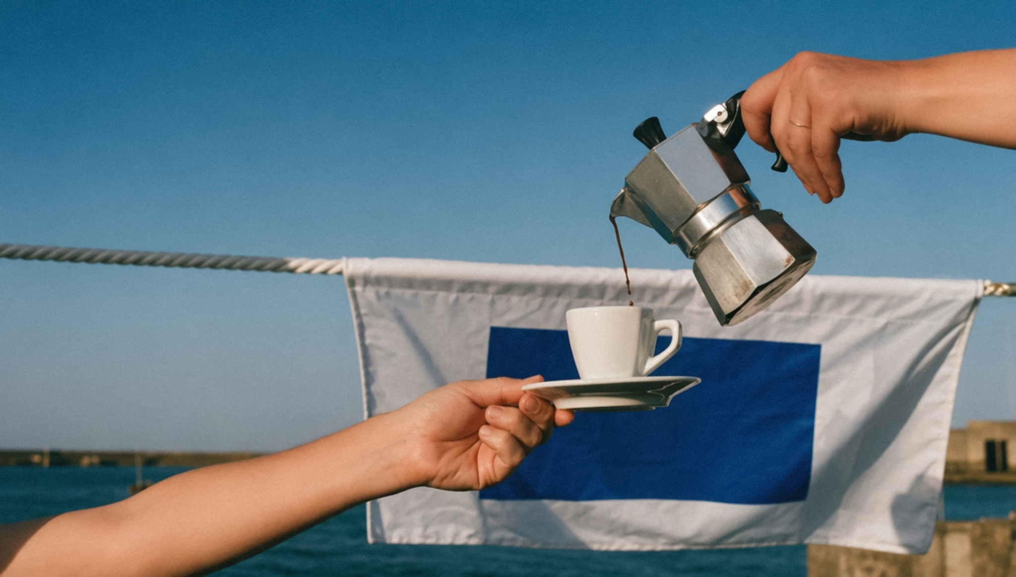 A hand pours espresso from a stovetop moka pot into a small white cup held on a saucer, set against a blue and white nautical flag, the sea, and a stone harbor wall under a bright sky.